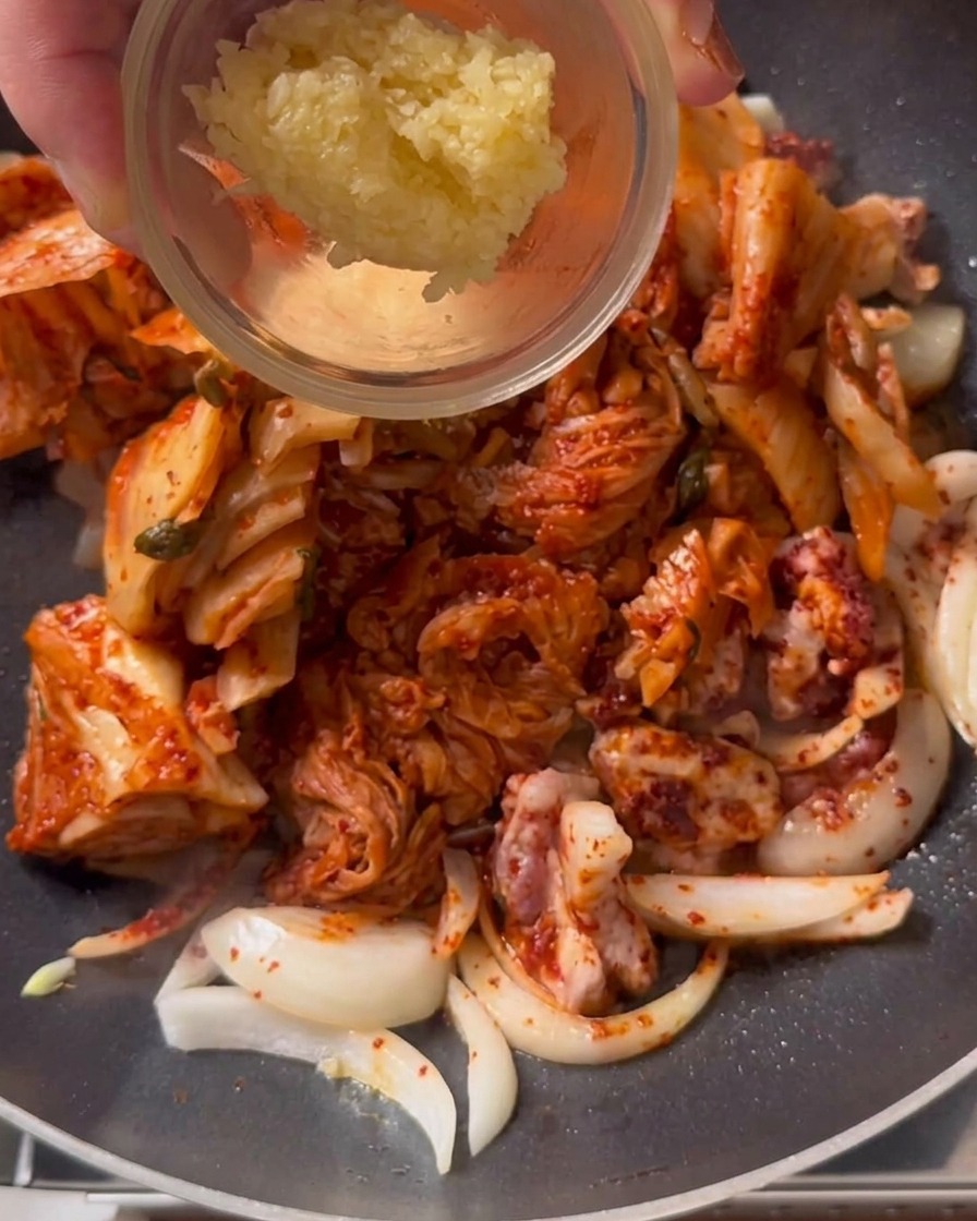 Una cucharada de ajo picado siendo añadida a una olla con kimchi y cerdo sofriéndose.