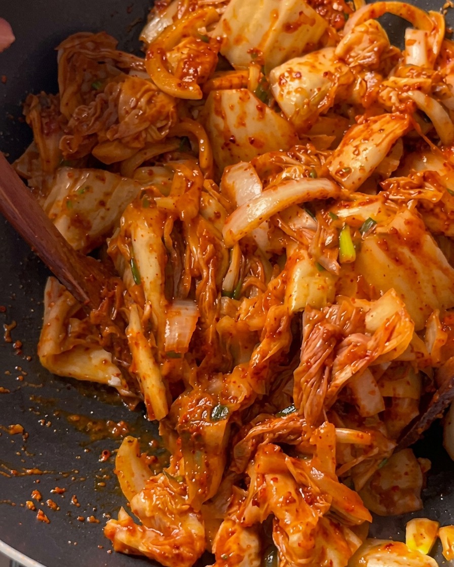 Trozos de kimchi siendo sofritos junto con cerdo y cebollas en un wok negro profundo usando una espátula de madera.