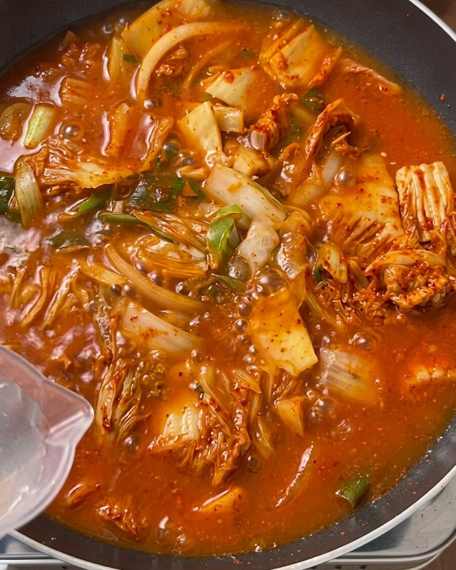 Agua limpia siendo vertida desde una jarra de plástico transparente en una olla grande llena de una mezcla burbujeante de kimchi y carne.