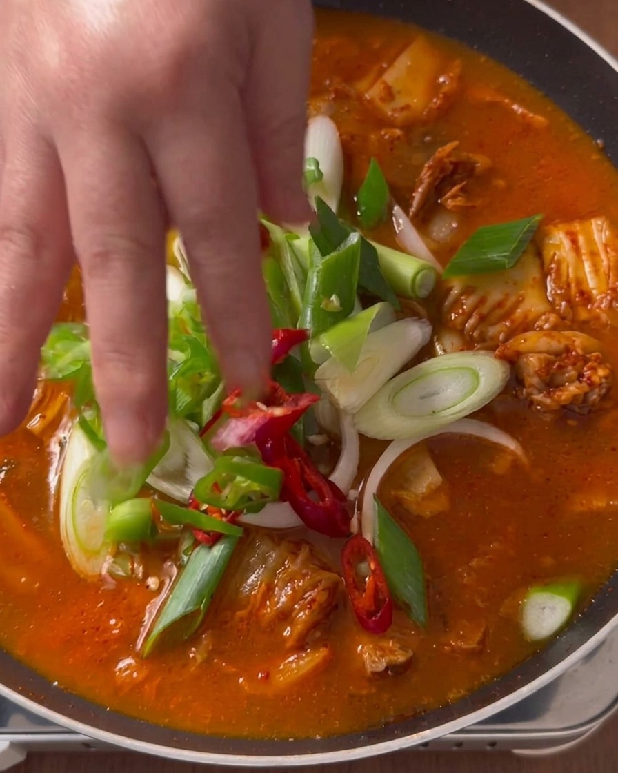 Una mano colocando cebollas de verdeo frescas, chiles verdes y rodajas de chile rojo sobre un estofado de kimchi hirviendo.