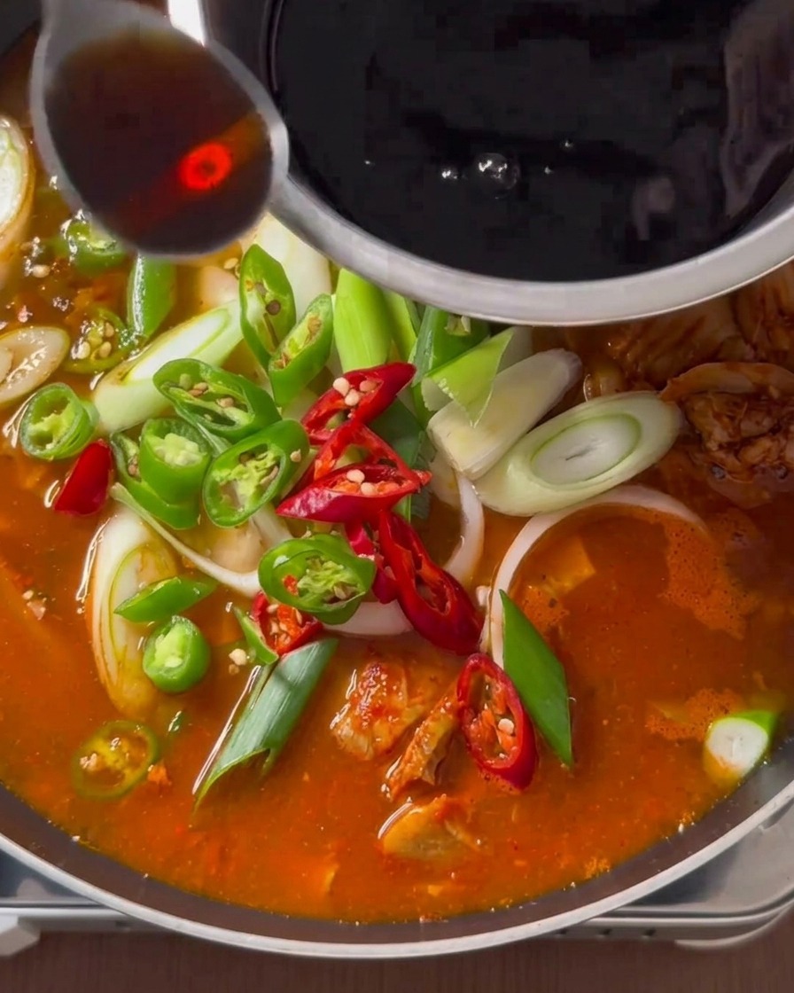 Una cucharada de sazón líquido oscuro siendo vertida desde un cuenco de acero inoxidable sobre un estofado de kimchi hirviendo.