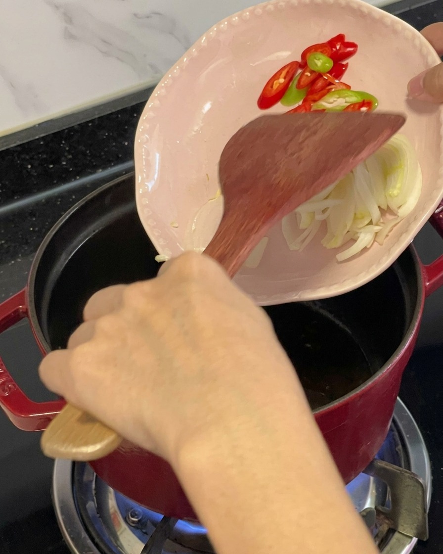Rodajas de chiles rojos y verdes con aromáticos siendo deslizados desde un plato rosa hacia una olla roja con aceite usando una espátula de madera.