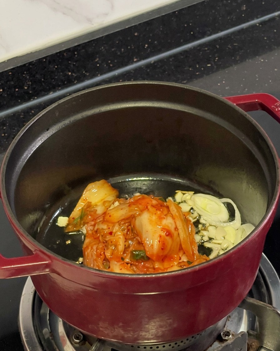 Kimchi rojo picado, ajo rebanado y cebollines cocinándose en aceite dentro de una olla.