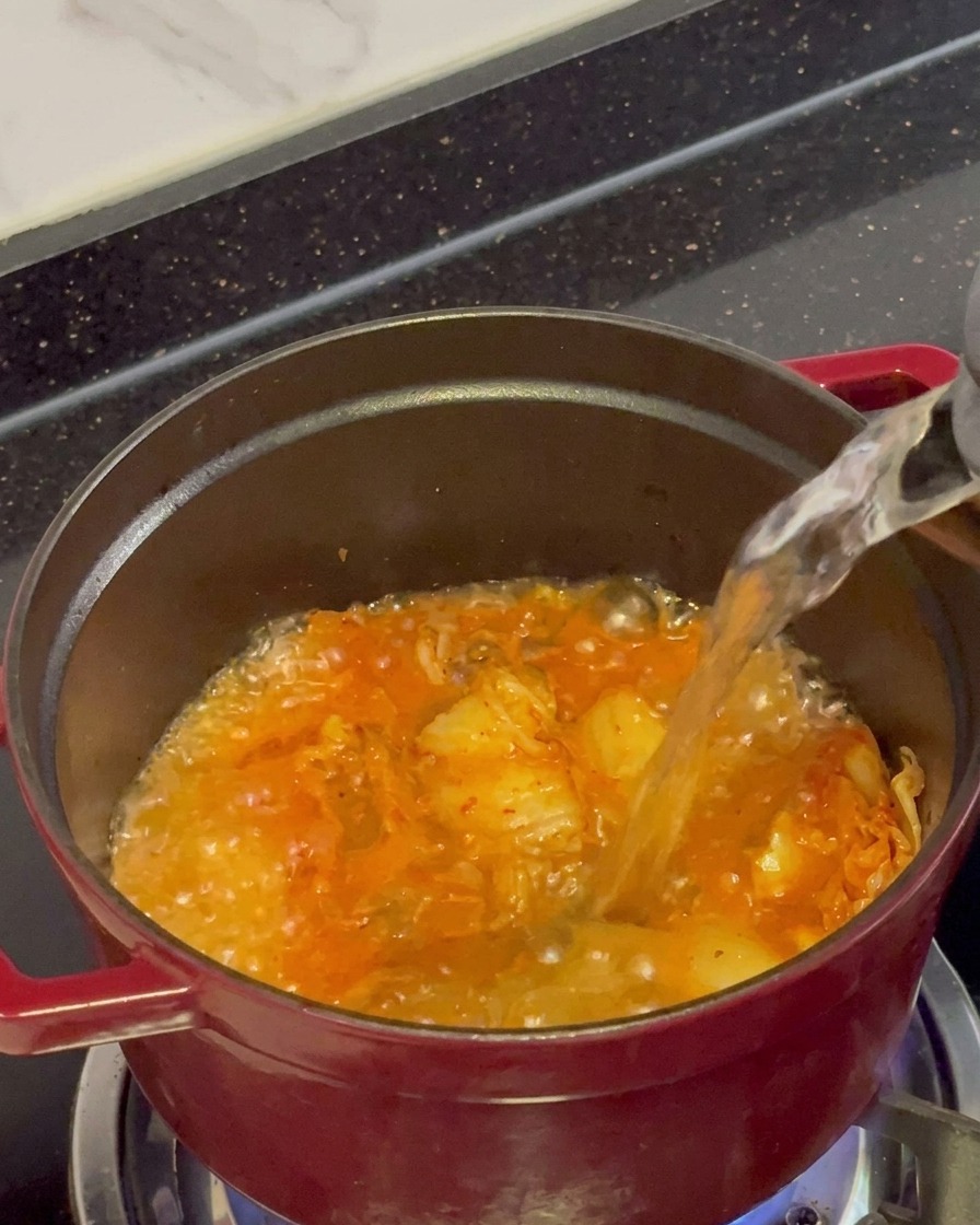 Agua hirviendo clara siendo vertida desde una tetera en una olla roja llena de kimchi salteado y burbujeante.