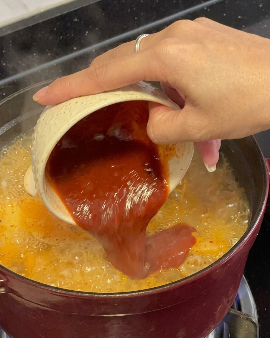 Una mezcla espesa de condimento de chile rojo oscuro siendo vertida desde un pequeño tazón jaspeado directamente en un estofado de mariscos rojo y burbujeante.