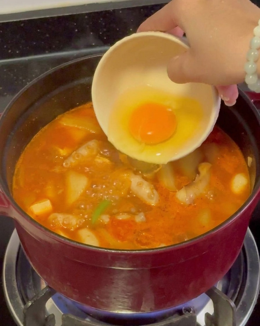 Un huevo crudo con una yema amarilla brillante siendo vertido suavemente desde un tazón pequeño en un estofado de mariscos rojo que hierve vigorosamente.