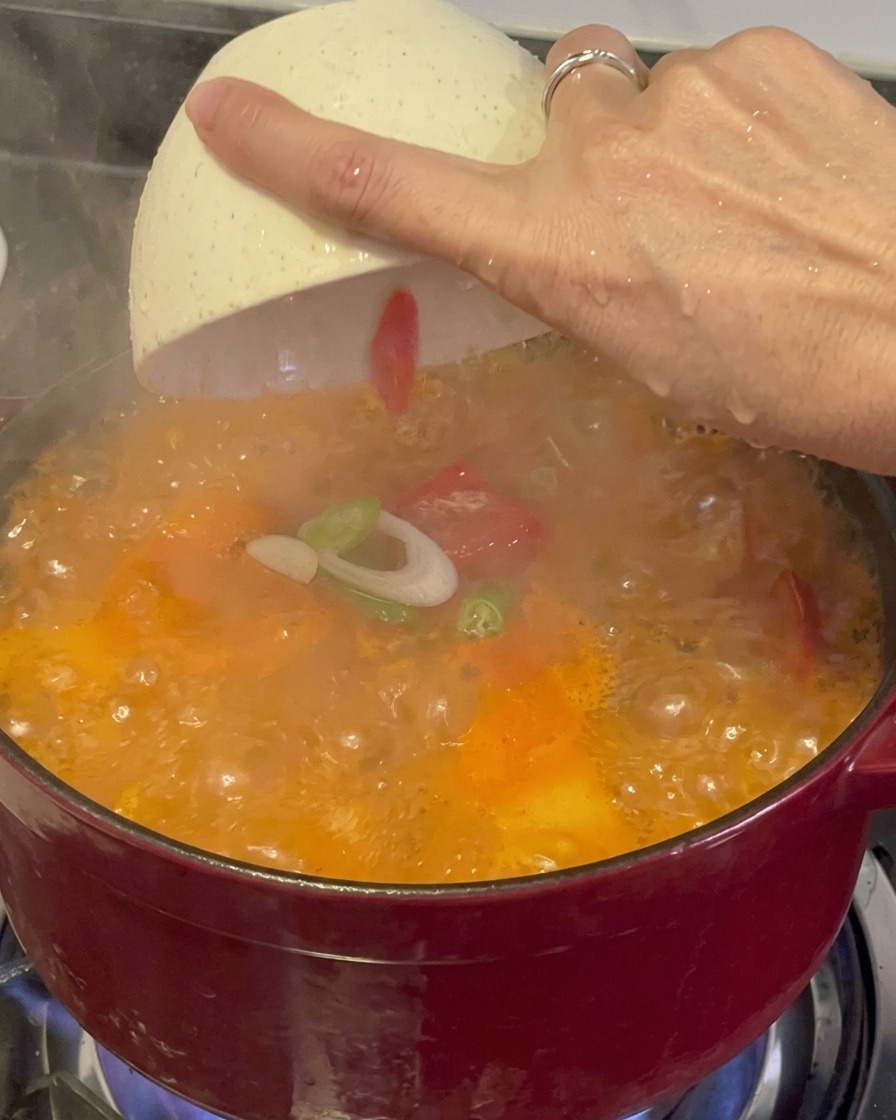 Cebollines verdes frescos picados y chiles rojos siendo espolvoreados desde un tazón blanco sobre un estofado de kimchi rojo, humeante y burbujeante.