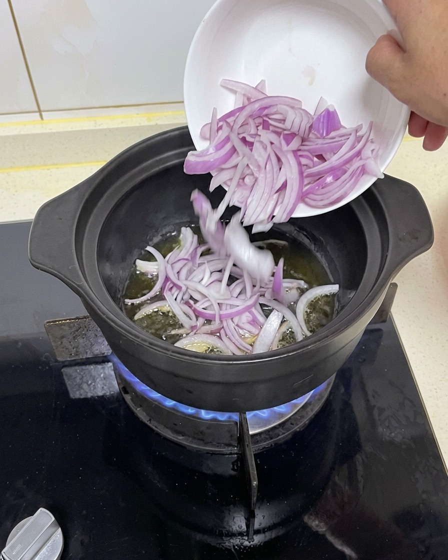 Rodajas de cebolla morada pasando de un pequeño cuenco blanco a una cazuela negra caliente con aceite sobre una llama de gas azul.
