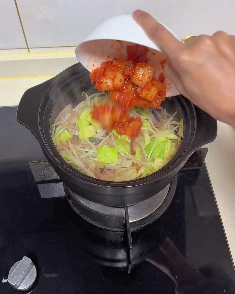 Una mano inclinando un cuenco blanco para añadir kimchi troceado de color rojo brillante en una cazuela de cerámica negra llena de cebollas traslúcidas, repollo verde y brotes de soja.