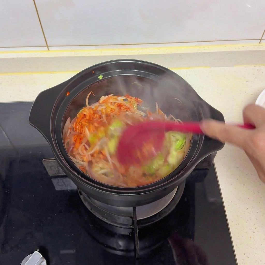 Salteando verduras mixtas y kimchi en una cazuela negra con una espátula roja para integrar la sal, la salsa de soja y los condimentos.