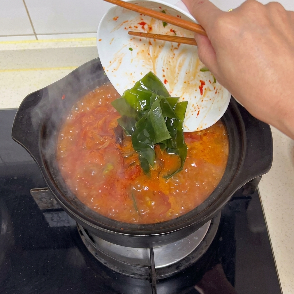 Unos palillos deslizando tiras anchas de alga kelp hidratada de color verde oscuro desde un plato blanco a una base de sopa sabrosa y roja en ebullición.