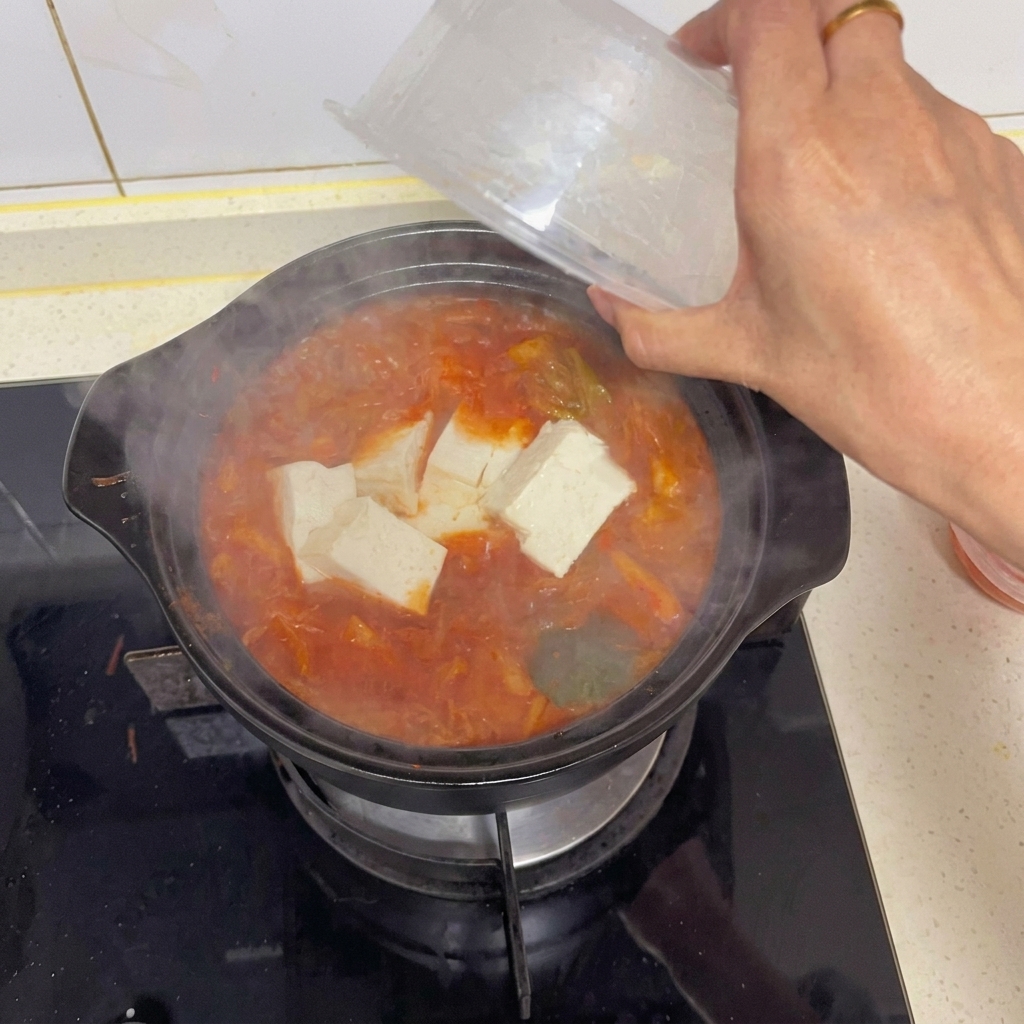 Una mano añadiendo delicadamente grandes cubos de tofu blanco desde un recipiente de plástico transparente a una cazuela de sopa de kimchi hirviendo.