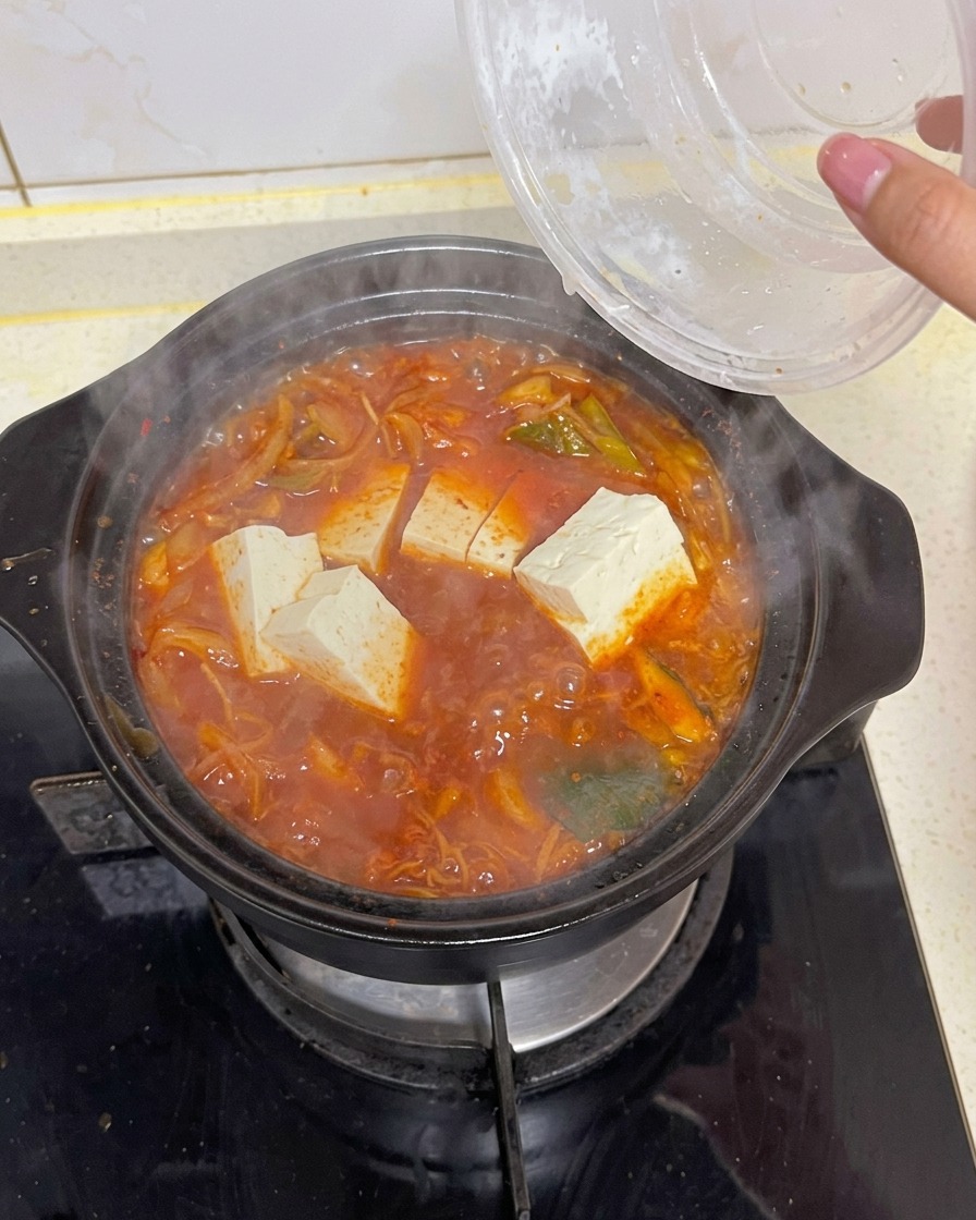 Bloques de tofu blanco rectangular cayendo desde un recipiente transparente a una cazuela burbujeante de sopa de kimchi roja con verduras y alga kelp.