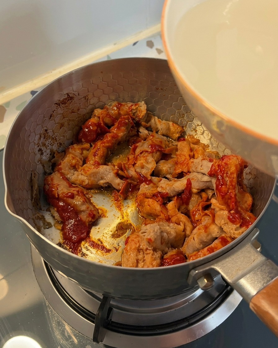 Agua turbia del lavado del arroz vertiéndose desde un tazón blanco a una olla con carne sazonada.