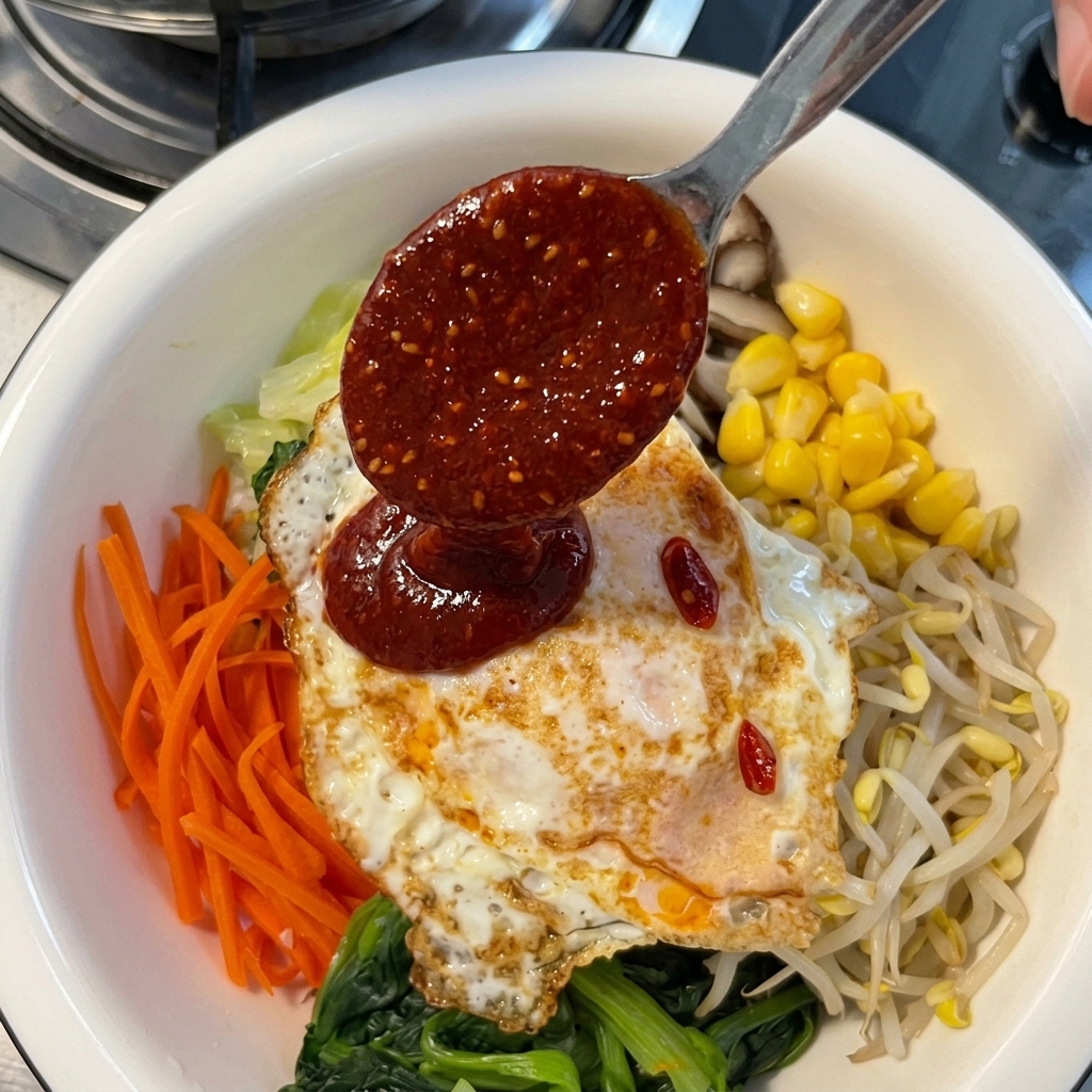 Vertiendo una salsa espesa de chile rojo desde una cuchara de metal sobre un huevo frito y vegetales de bibimbap de varios colores.