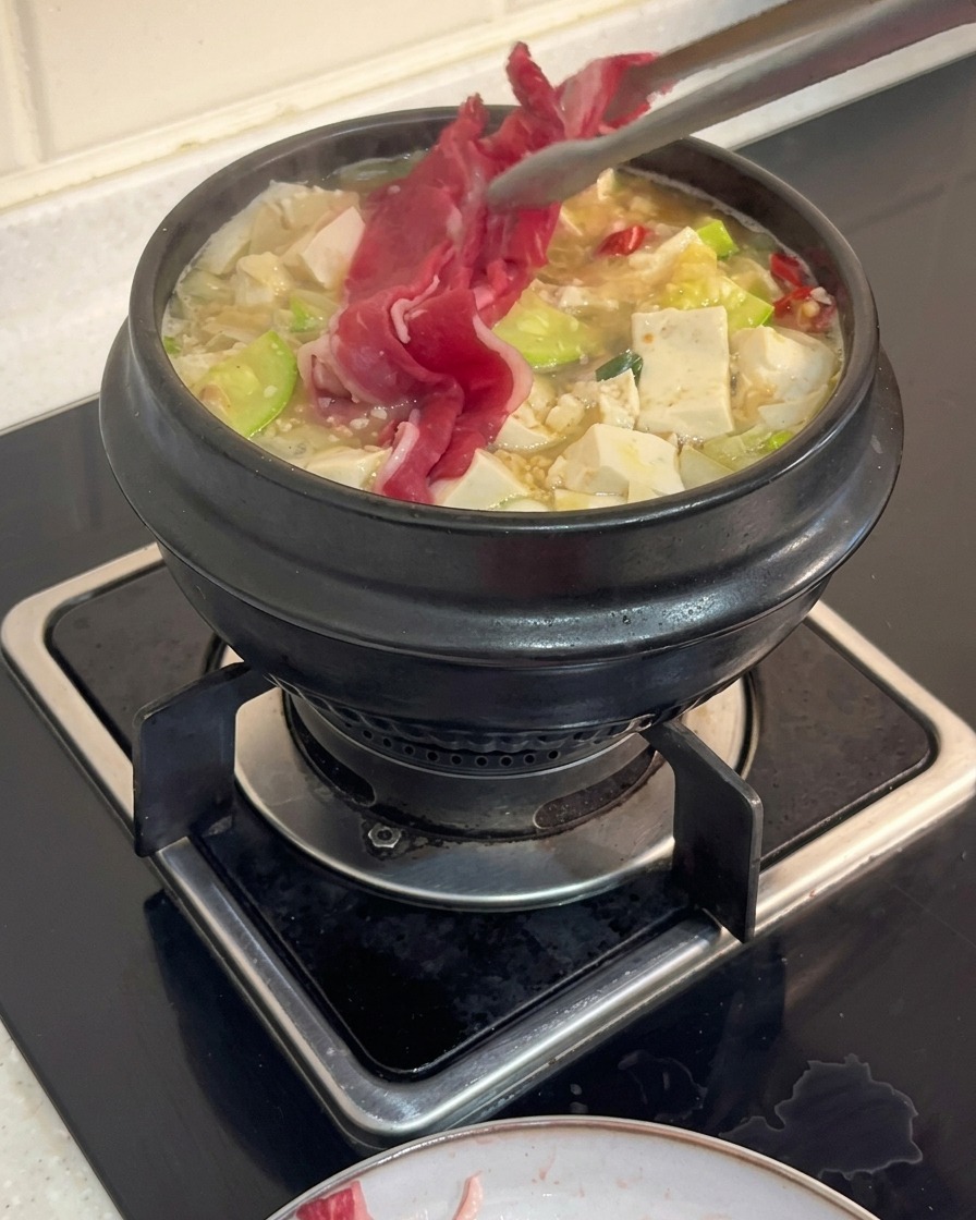 Unas pinzas de cocina colocando finas láminas de carne de res cruda en una cazuela hirviendo de estofado coreano lleno de tofu y calabacín.