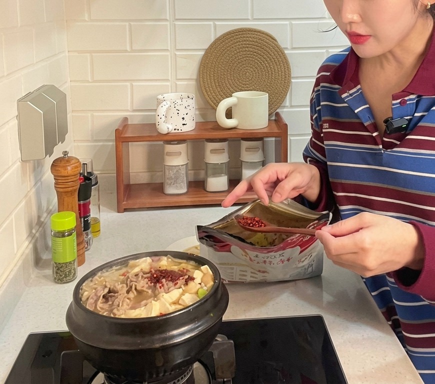 Una cucharada de chile rojo en polvo grueso siendo añadida a una cazuela de barro hirviendo con estofado coreano de pasta de soja lleno de tofu y carne de res en láminas.