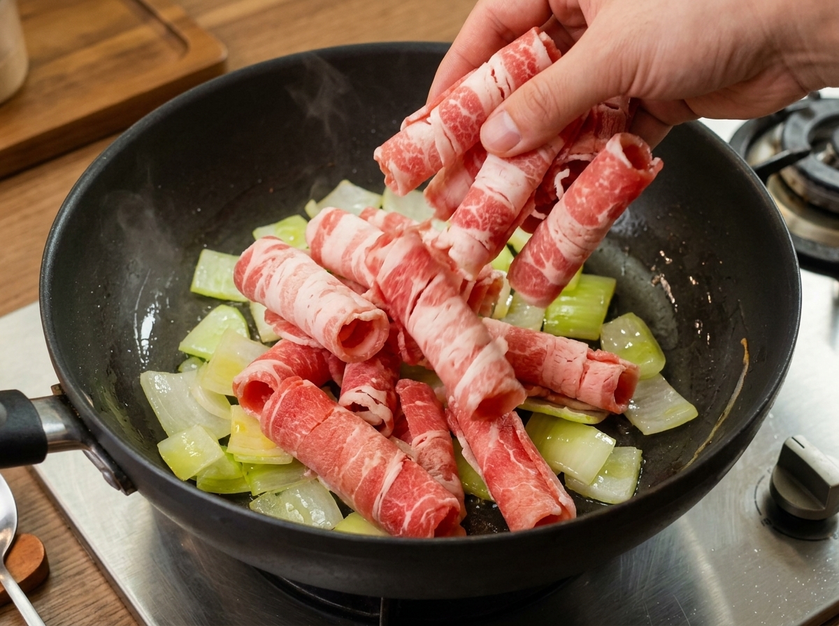 Láminas finas de ternera cruda siendo colocadas en una olla con cebolla salteada translúcida.