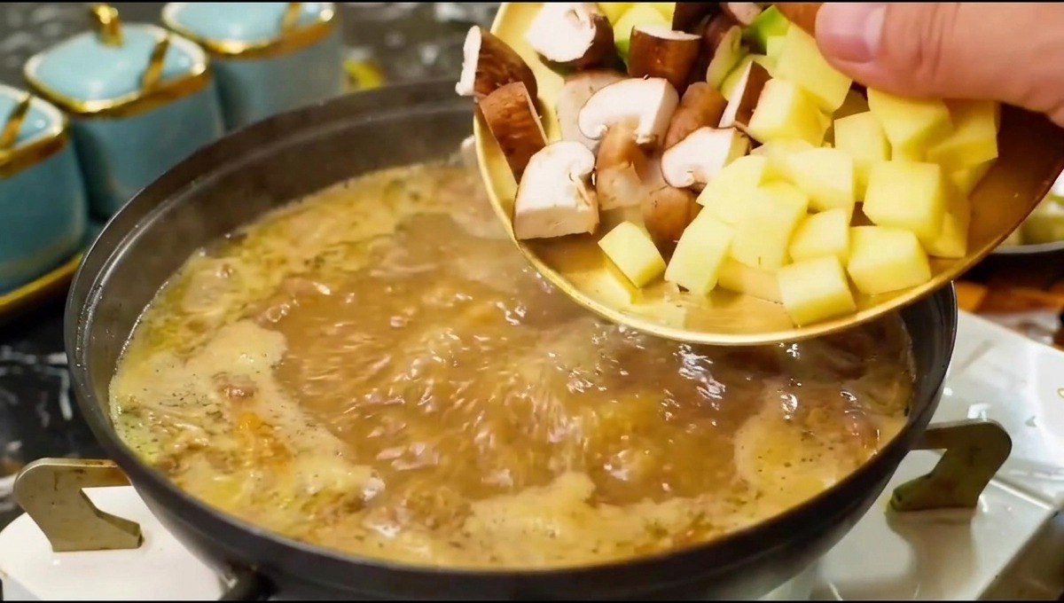 Setas troceadas y verduras deslizándose desde un plato hacia una olla con caldo sazonado hirviendo.