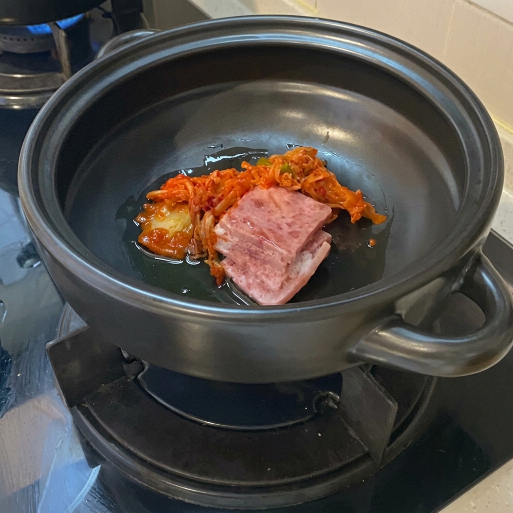 Rebanadas de panceta de cerdo cruda y kimchi picado vibrante reposando en aceite en el fondo de una olla de barro cerámica oscura.
