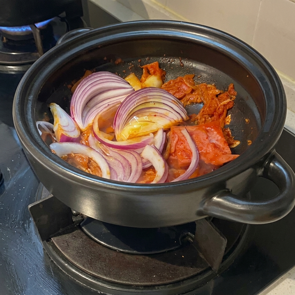 Rebanadas gruesas de cebolla roja vibrante dispuestas sobre un salteado sazonado de kimchi y panceta de cerdo dentro de una olla de barro.