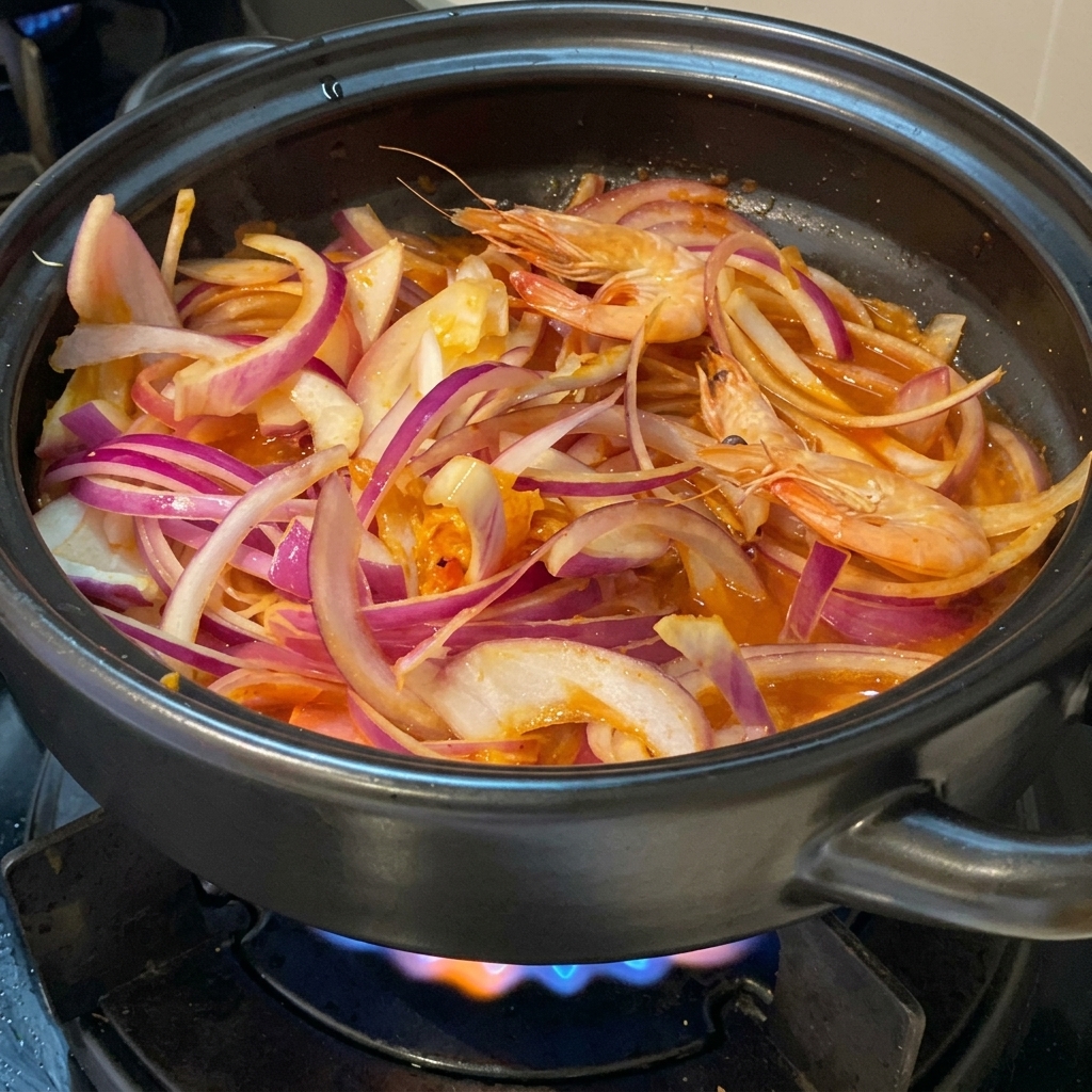 Camarones enteros rosados y frescos acomodados entre cebollas rojas rebanadas y kimchi en una olla de barro negra sobre una estufa a gas encendida.