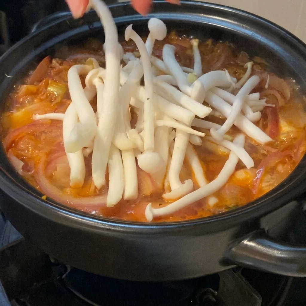 Setas de aguja blanca frescas siendo colocadas cuidadosamente a mano sobre una sopa de kimchi roja burbujeante dentro de una olla de barro negra.