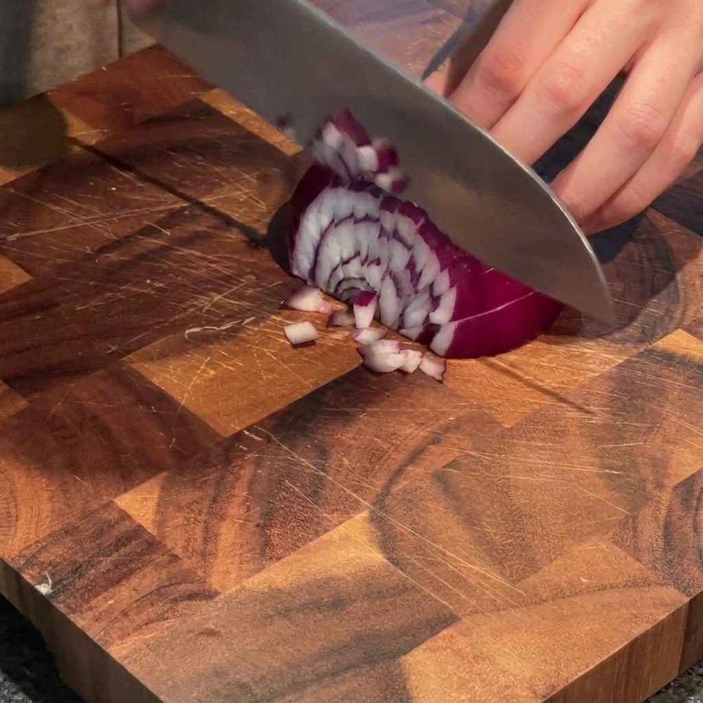 Una mano usando un cuchillo de chef para picar una cebolla morada en trozos pequeños sobre una tabla de cortar de madera.