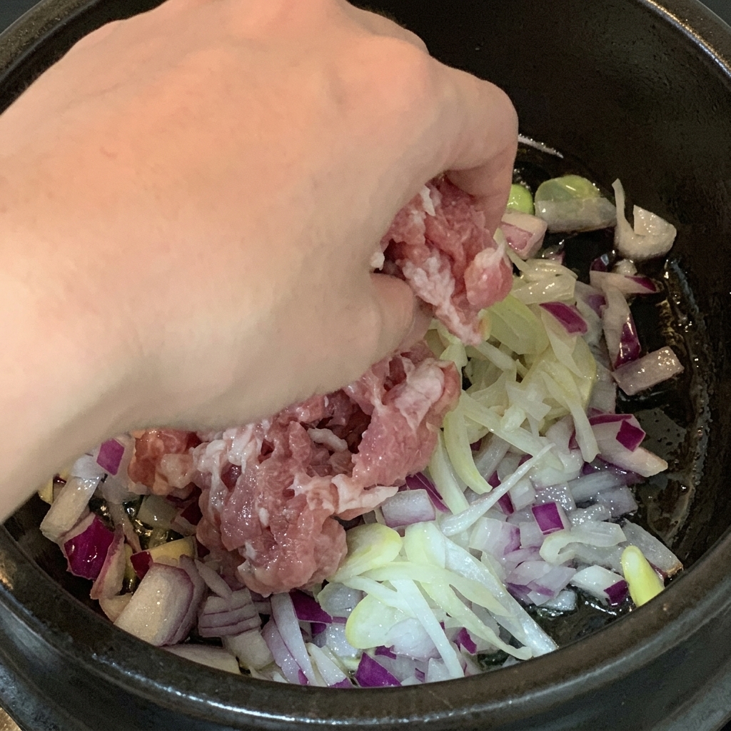 Trozos de panceta de cerdo cruda añadiéndose a una olla que contiene cebolla morada picada y cebolletas en tiras.