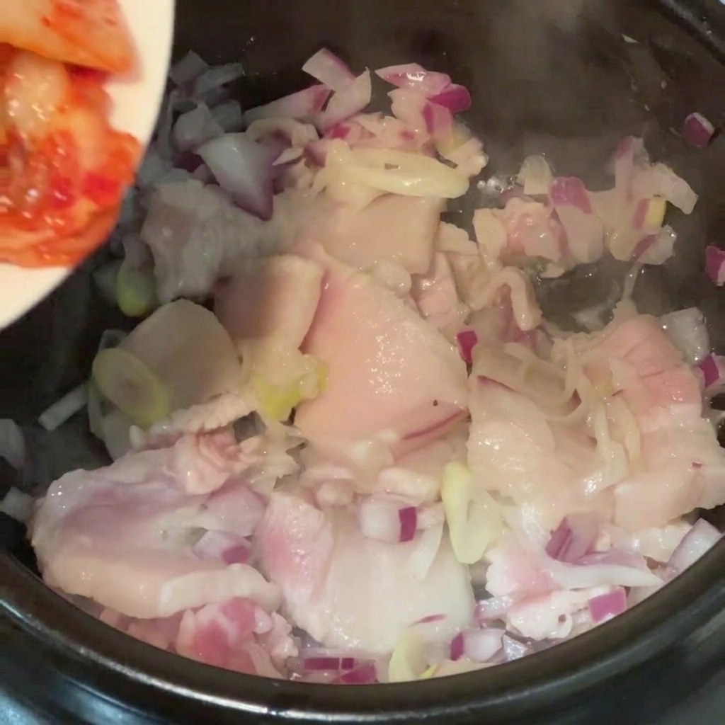 Col de kimchi picada volcándose desde un plato a una olla con trozos de panceta de cerdo y cebolla morada.