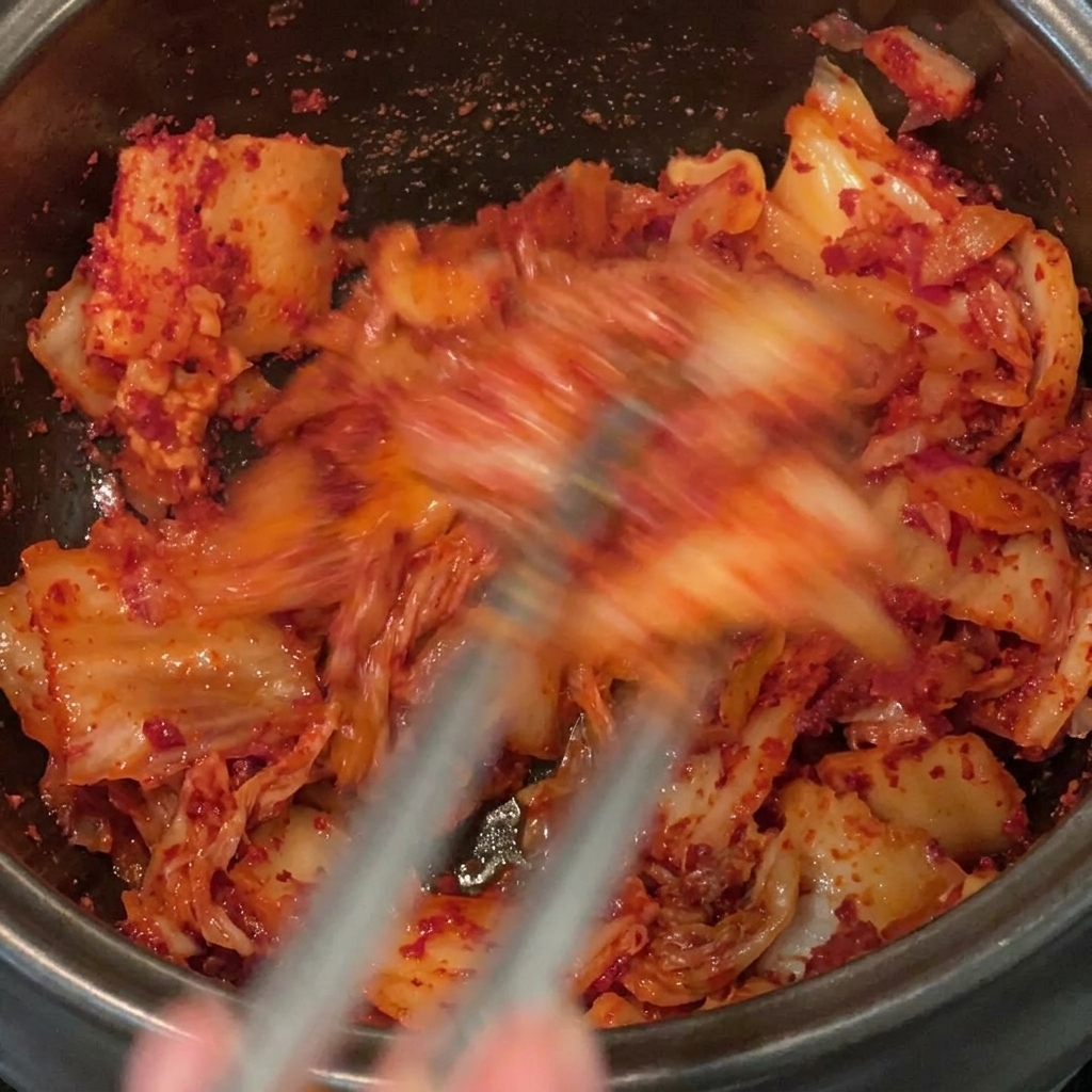 Removiendo y mezclando bien el chile en polvo con el kimchi troceado y la panceta dentro de una olla negra.