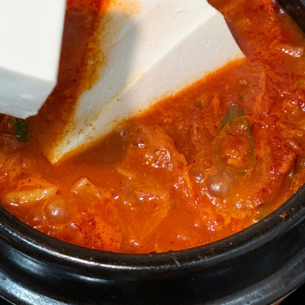 Un bloque grande de tofu sedoso blanco colocándose cuidadosamente en una olla hirviendo de sopa de kimchi roja.