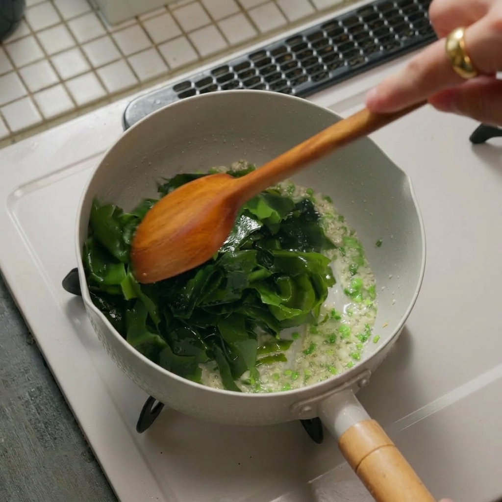 Montón de algas rehidratadas de color verde vibrante que se añaden a una olla con los aromáticos, siendo removidas por una espátula de madera.