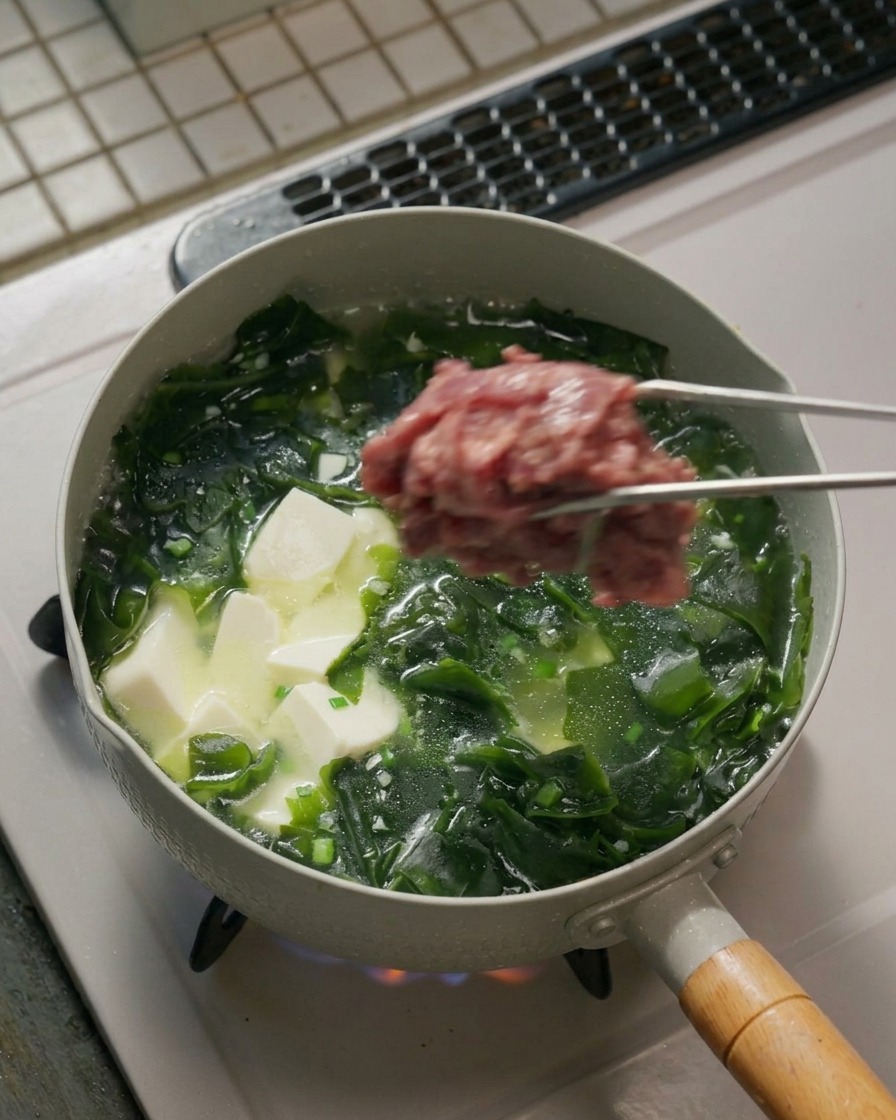 Pinzas de metal sujetando un grupo de rodajas de carne marinada cruda sobre una olla hirviendo llena de sopa de algas y tofu.