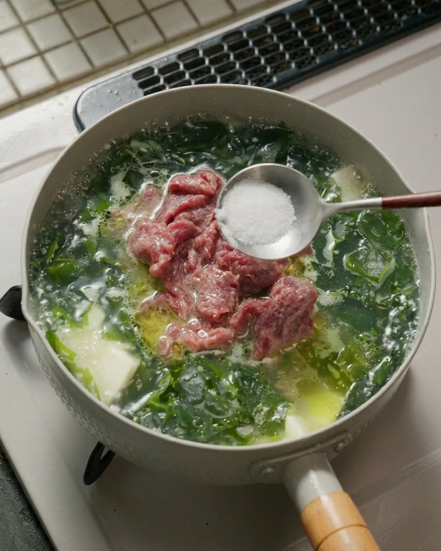 Cuchara de metal con sal blanca y pimienta en polvo sobre una olla de sopa de algas a fuego lento.