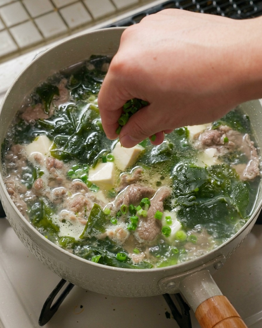 Mano espolvoreando una generosa pizca de cebolleta finamente picada sobre una sartén caliente con sopa de algas a fuego lento.