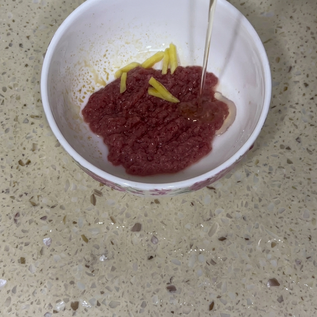Carne de ternera picada cruda en un cuenco de cerámica blanca con láminas de jengibre y un chorrito de aceite de cocina siendo vertido.