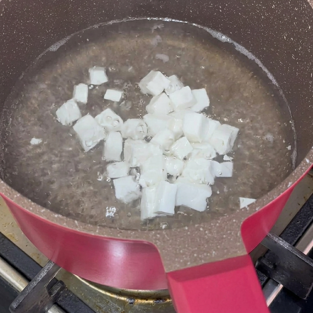 Pequeños cubos de tofu blanco suave hirviendo en agua dentro de una olla rosa.