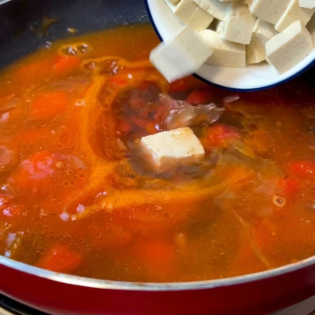 Cubos blancos de tofu tierno cayendo desde un pequeño bol en una sartén roja llena de una base de sopa de tomate burbujeante.
