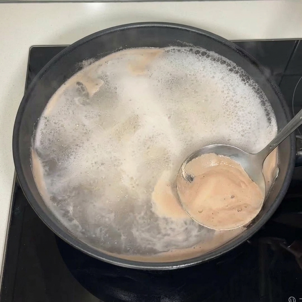 Un cucharón metálico retirando la espuma marrón y las impurezas de la superficie del caldo hirviendo.