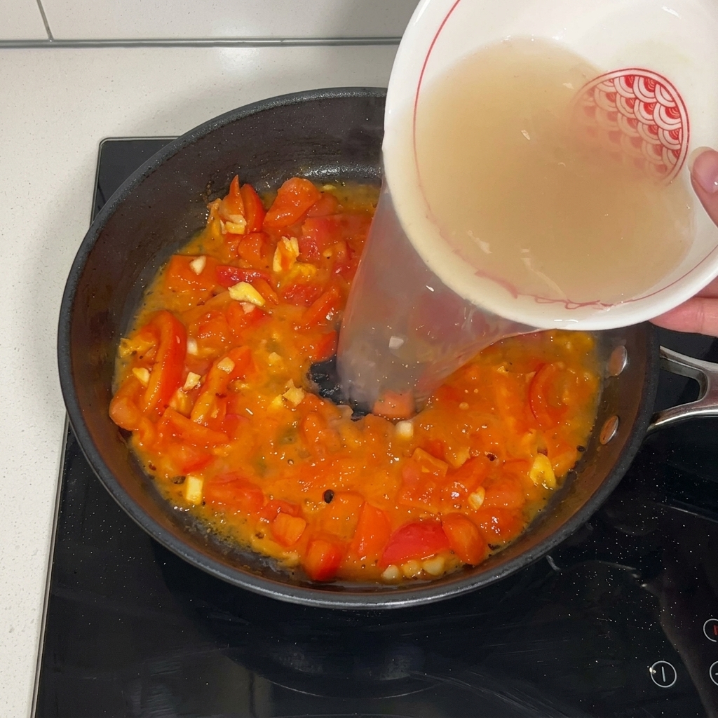 Caldo de ternera vertiéndose desde un cuenco de cerámica blanca sobre los tomates sofritos en la sartén.