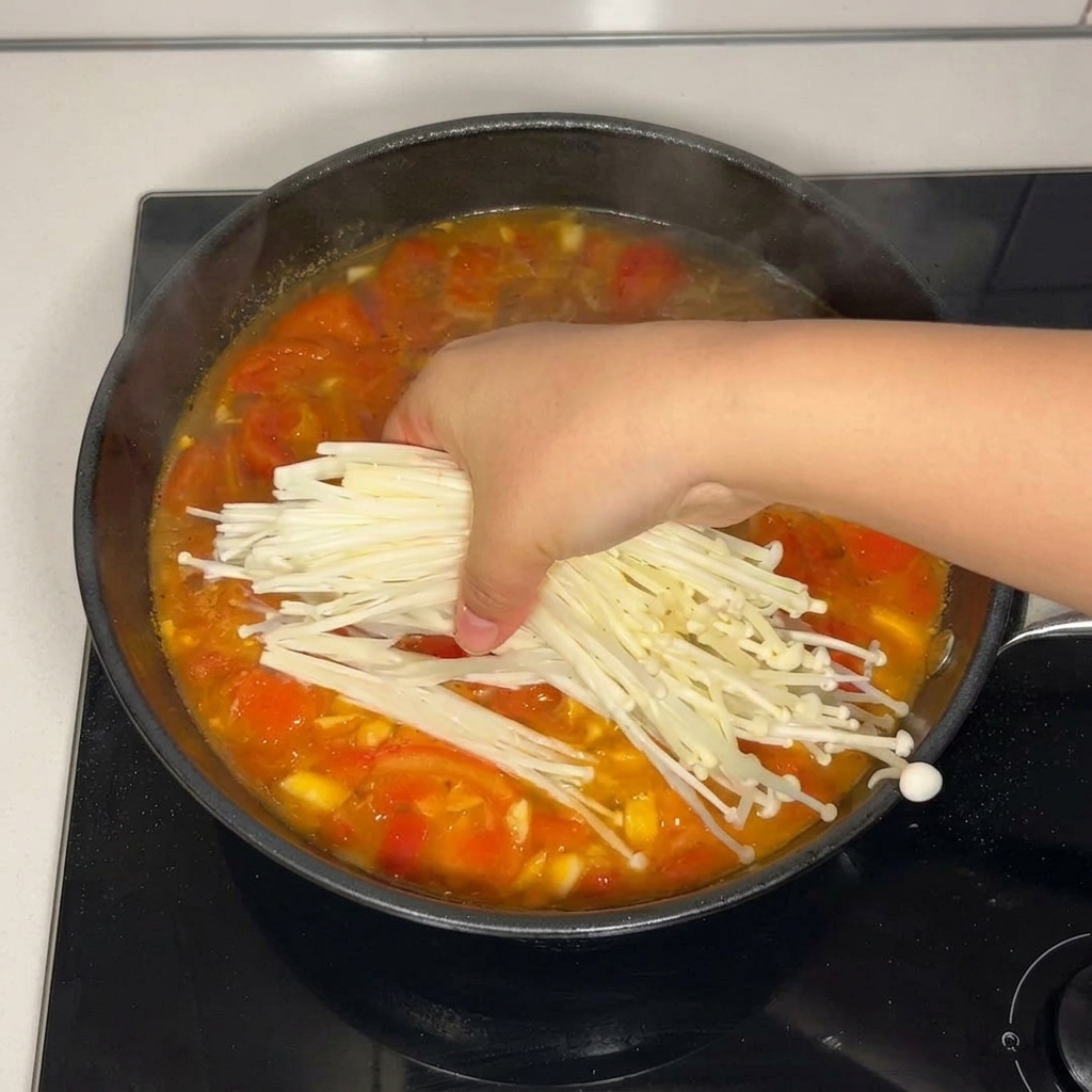 Una mano añadiendo un manojo de setas enoki frescas a la base de sopa de tomate.