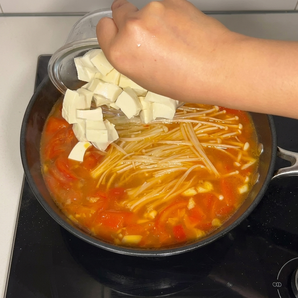 Cubos de tofu suave siendo vertidos desde un cuenco de cristal a la sopa de tomate y setas.