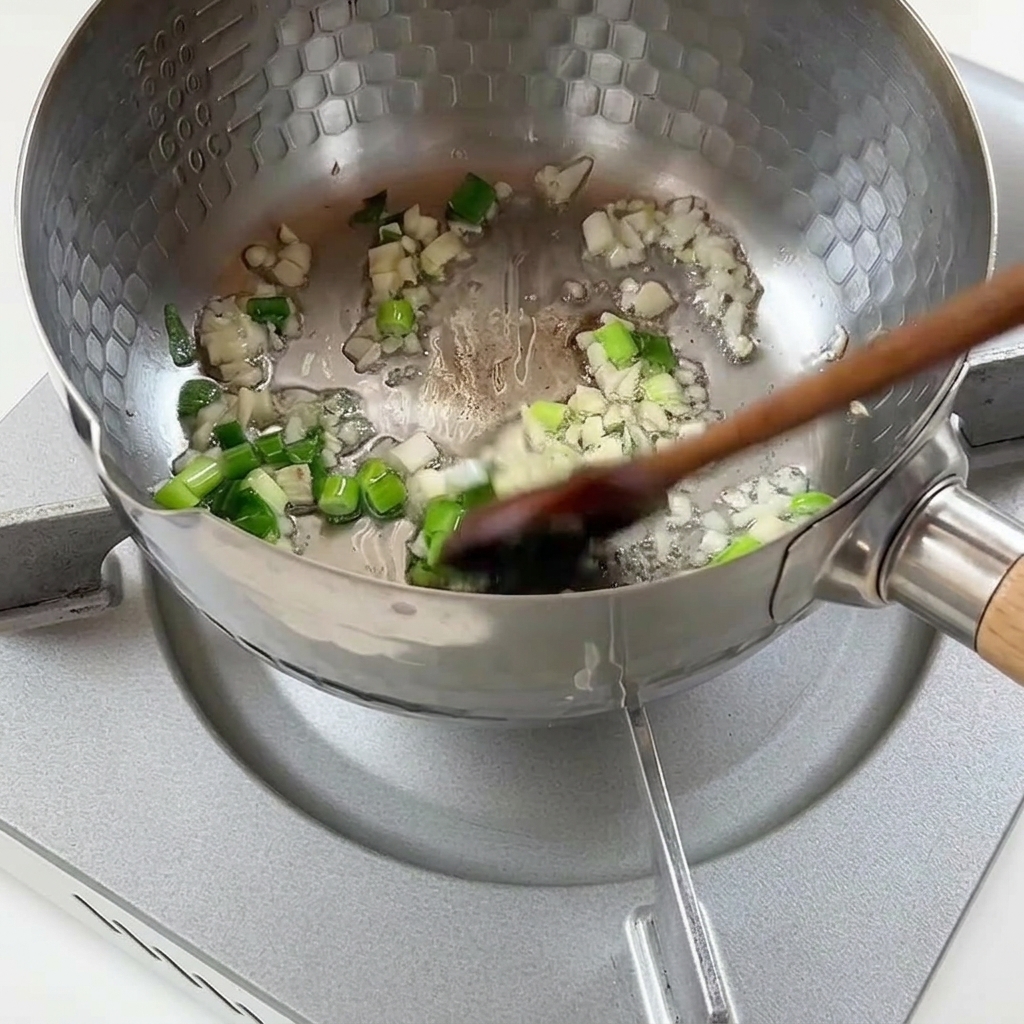 Cebolleta picada y ajo picado friéndose en aceite caliente dentro de una olla de metal.