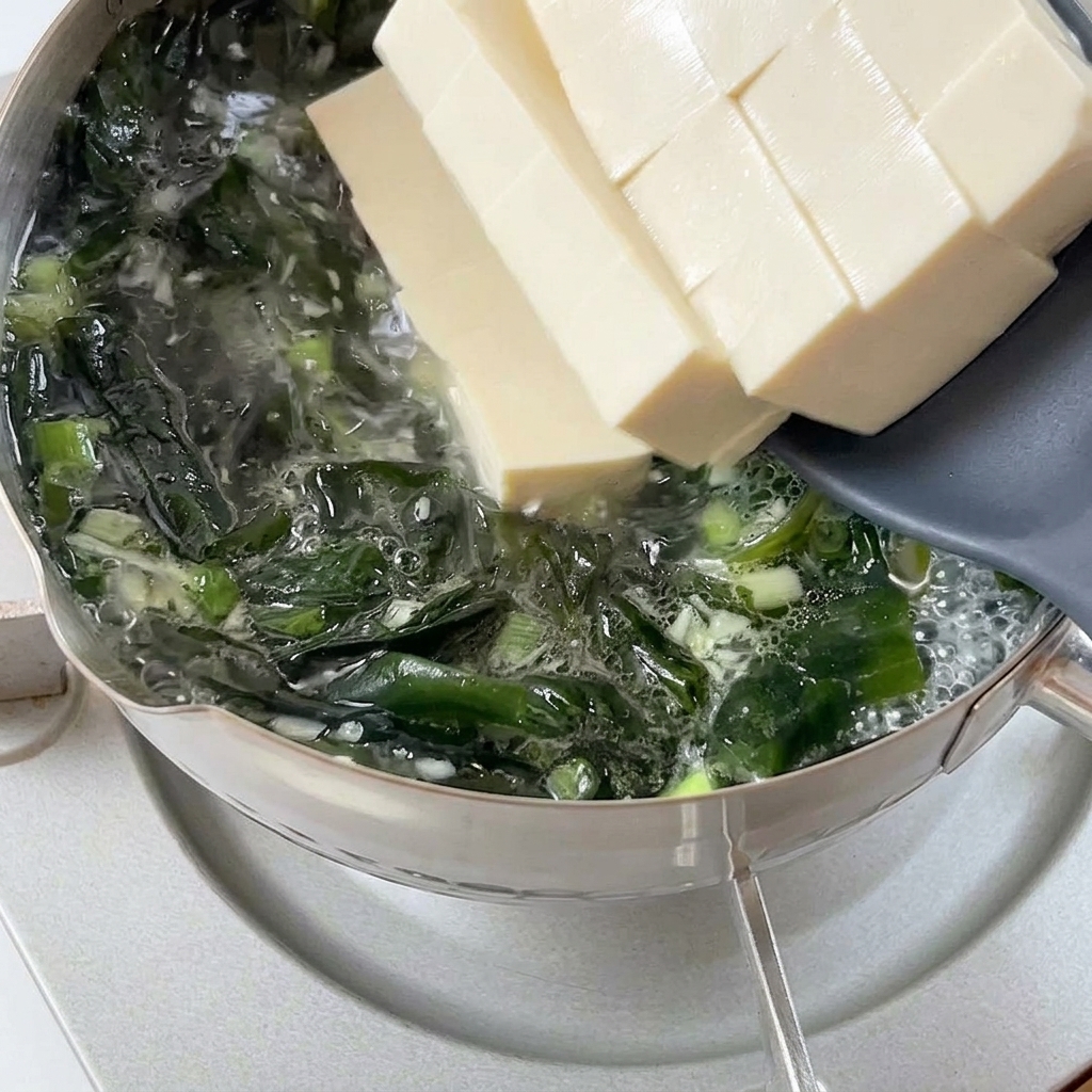 Gruesas rodajas de tofu blanco suave deslizándose desde una espátula gris a una olla burbujeante con caldo de wakame.