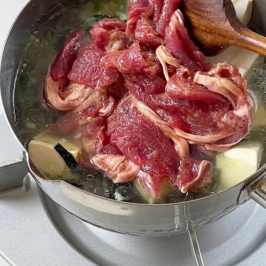 Rodajas de ternera cruda marinada introduciéndose suavemente en una olla caliente de sopa de wakame y tofu.