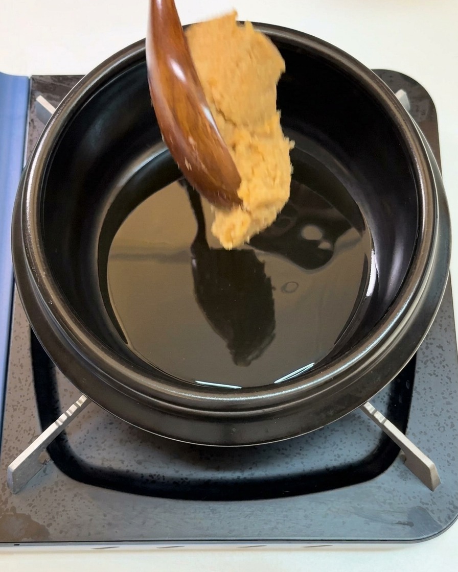 Una cucharada grande de pasta de soja espesa cayendo de una cuchara de madera a una olla negra con aceite de sésamo.