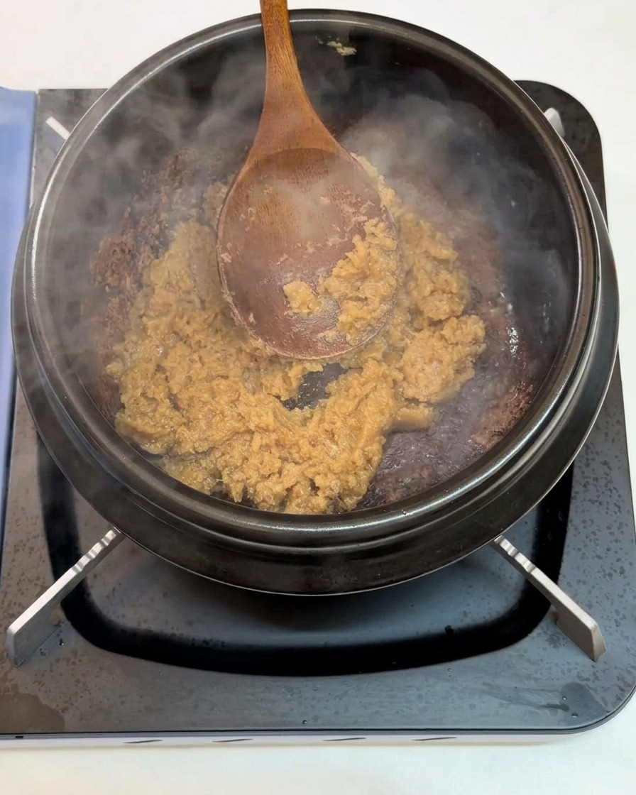 Una cuchara de madera sofriendo ajo picado y pasta de soja dentro de una olla de barro negra al fuego.