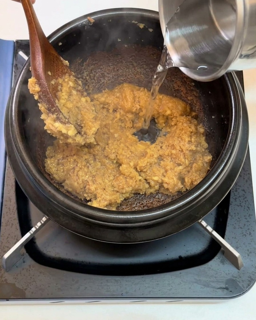 Agua cayendo desde un recipiente metálico hacia una olla negra que contiene la base de pasta de soja sofrita.