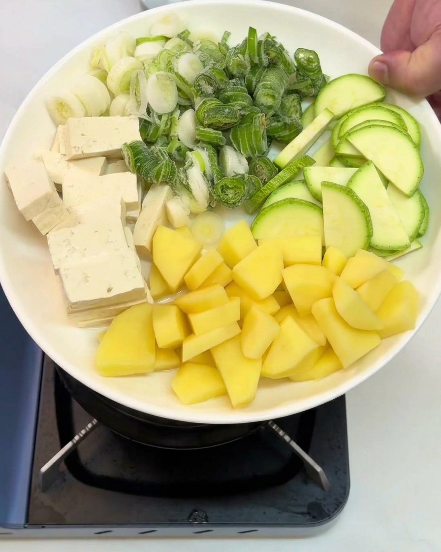 Un plato blanco sostenido sobre una estufa con secciones de tofu en cubos, cebolletas picadas, calabacín en rodajas y patatas crudas en dados.
